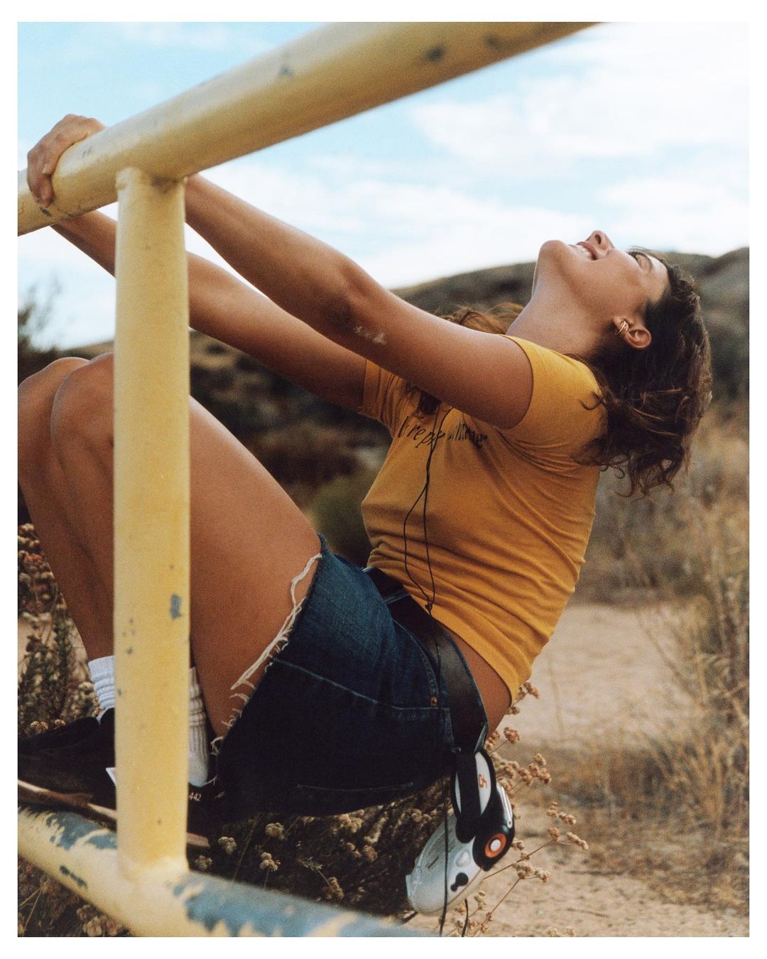 Woman in denim shorts and a yellow t-shirt is hanging from a yellow metal rail outside on a sunny day with a nature background.
