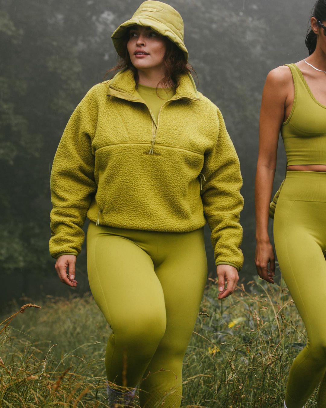 Two women walking in activewear. The first woman wears a chartreuse colored fleece, leggings, and bucket hat in an outdoor setting.