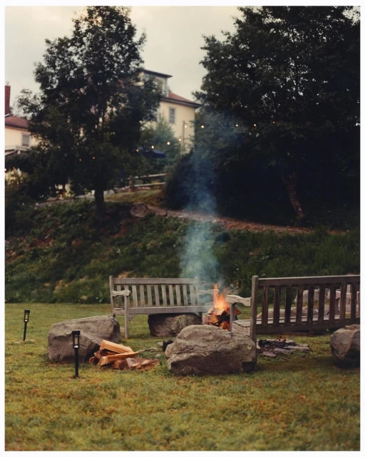 An outdoor fire pit with firewood and benches, surrounded by boulders on a grassy lawn, with trees and a building in the background.