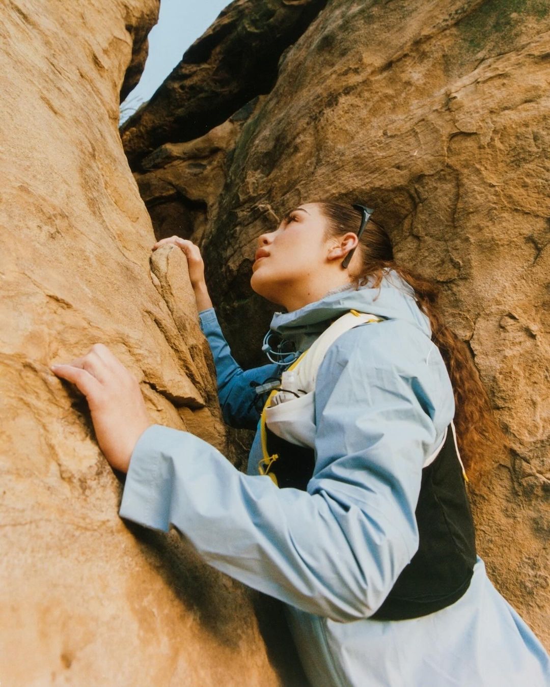 Woman rock climbing in a rugged, rocky environment, wearing a light blue jacket and black vest, looking up at the rock face.