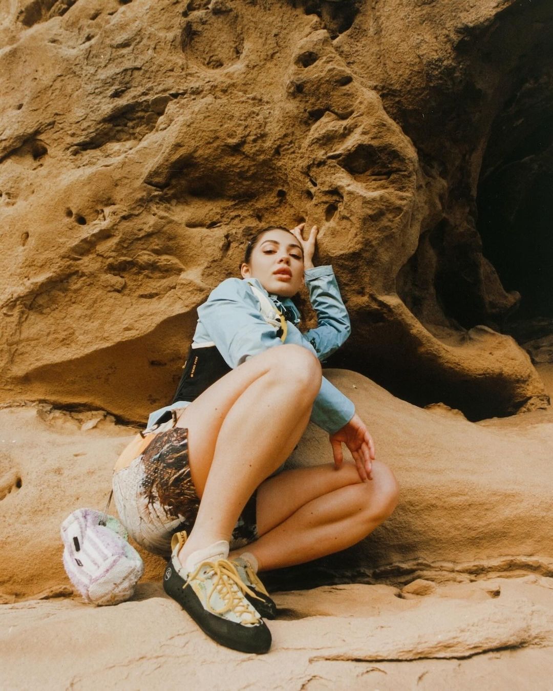 Young adult female in climbing shoes poses against a sandstone rock formation; earth tones, casual athletic wear, outdoor lifestyle.