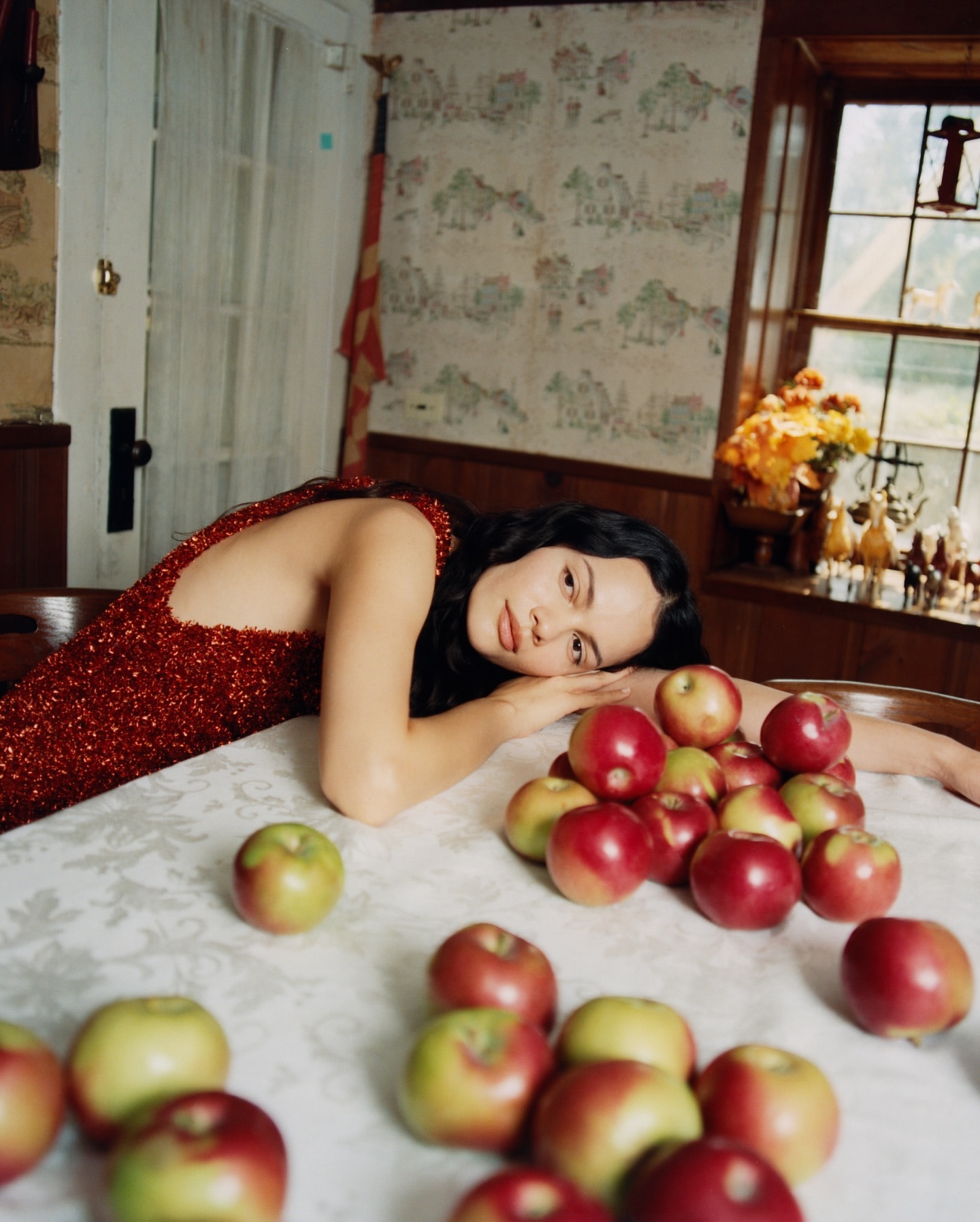 Woman in red dress poses at a table covered in red and green apples, indoor setting with patterned wallpaper, and window light.