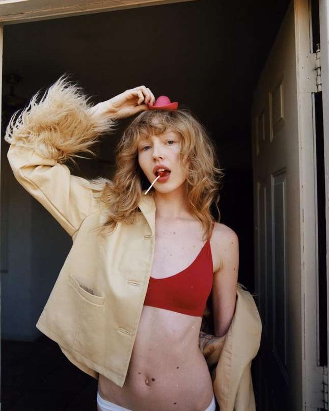 Blonde woman poses indoors with a red bralette, white underwear, jacket, and a red miniature hat holding a lollipop in her mouth.