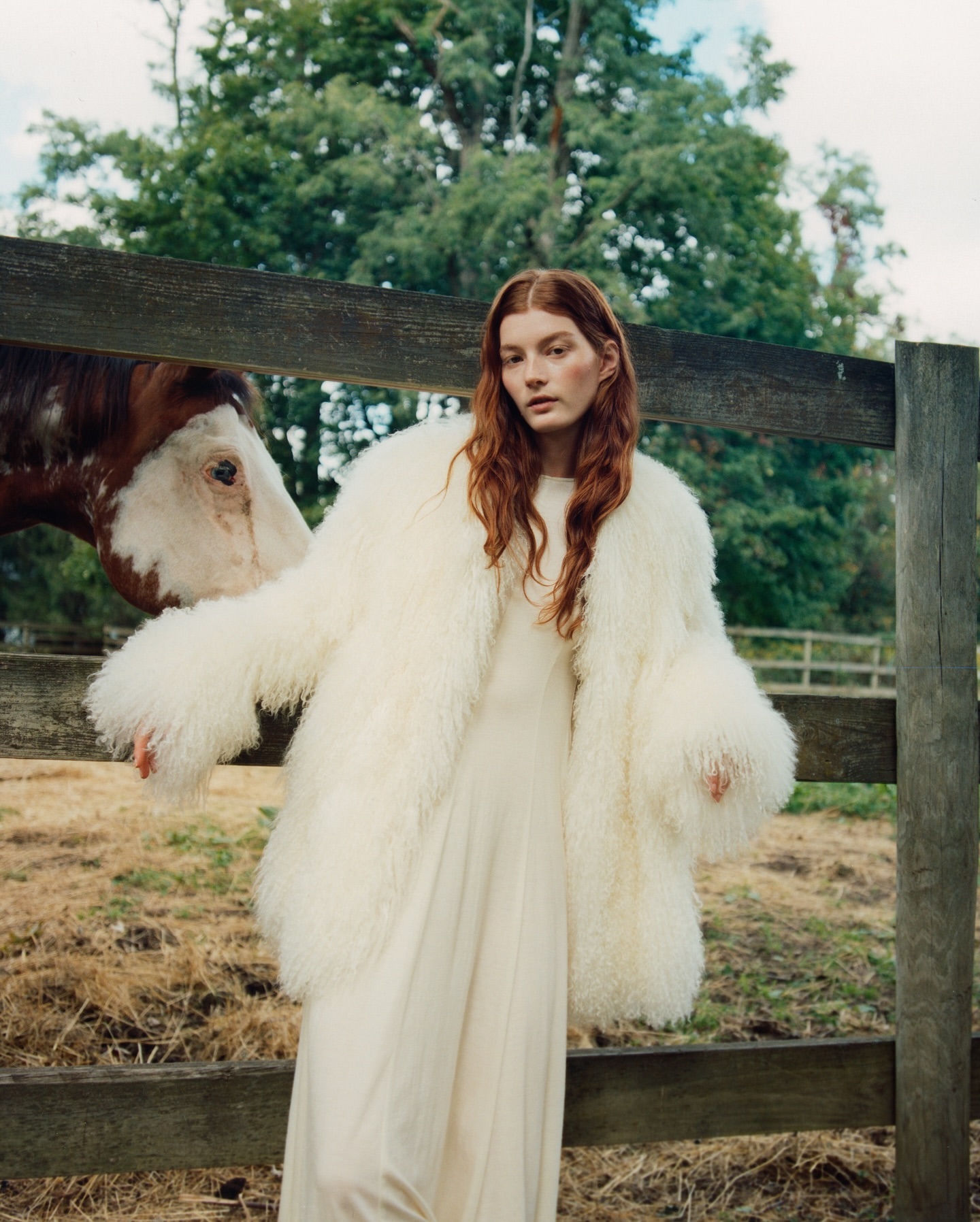 A stylish woman with red hair poses outdoors in a white dress and a fluffy white coat, standing near a rustic wooden fence.