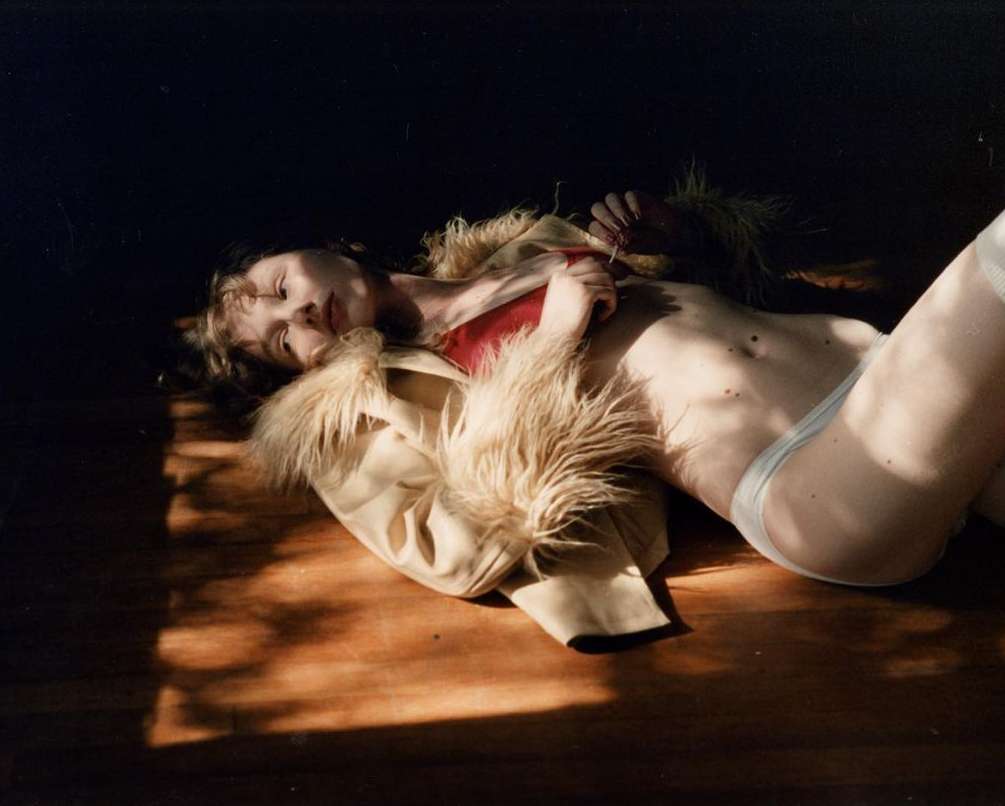 A woman lies on her side in a dimly lit room, her bare torso and legs illuminated by sunlight. She wears light underwear and a fur-trimmed jacket.