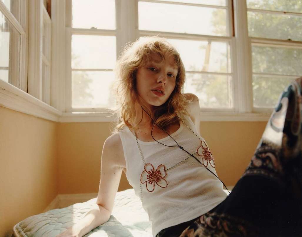 Blonde woman with white tank top poses indoors by sunlit window wearing floral necklace and patterned pants, looking at the camera.