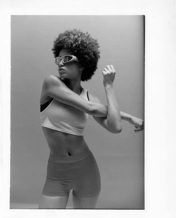 Black woman with afro and sunglasses stretching in athletic top and shorts against a neutral grey background