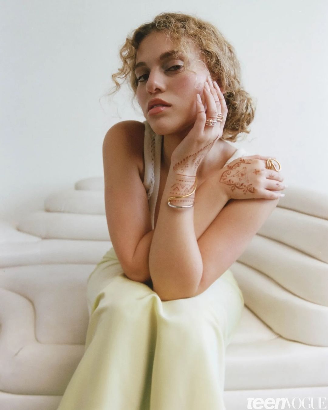 Portrait of a young woman with curly hair, wearing a light yellow dress and jewelry, posing in front of a white studio backdrop.