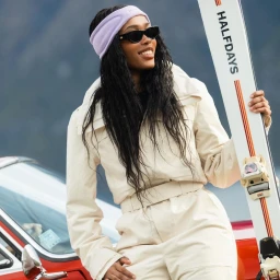 Black woman wearing a cream ski suit, headband, and sunglasses, holding a white ski with red details.