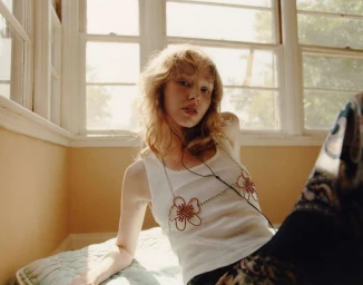 Young woman with blonde hair wearing a white tank top and beaded flower body chain seated indoors by a sunny window.