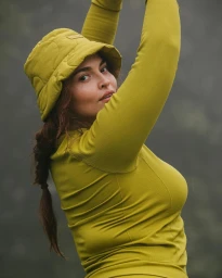 Woman wearing an olive green bucket hat and matching long-sleeve top with her arms raised against an outdoor backdrop.