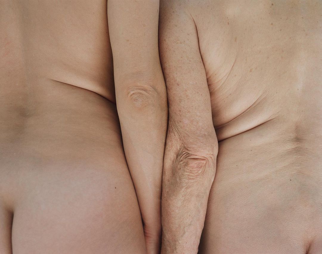 Close-up view of two nude human torsos, one fair and one elderly, pressed together. The left side is paler and smoother, while the right shows more wrinkles.