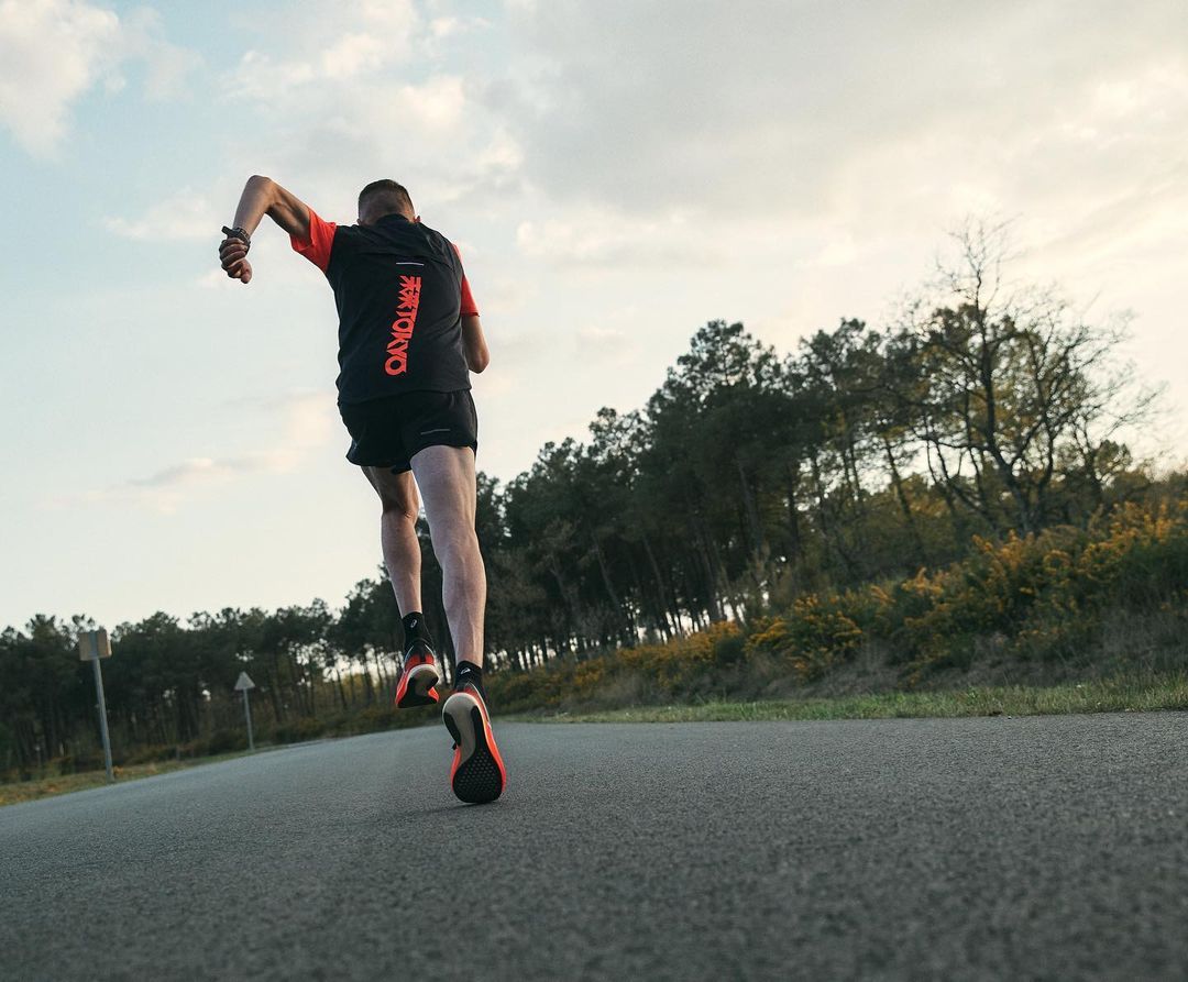 Athletic man running on asphalt road with a forest and cloudy sky. He wears dark athletic clothes, and the camera follows from behind.