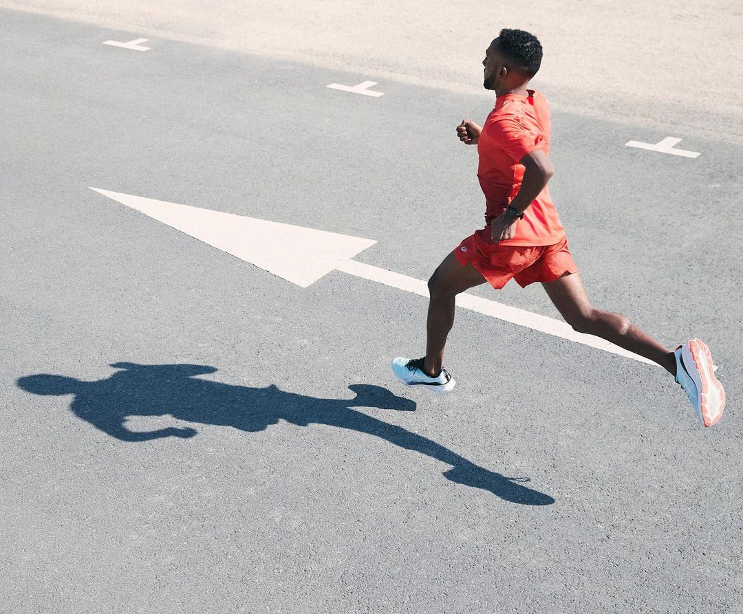 An athletic Black man in a red sports outfit runs on an asphalt road marked with an arrow, casting a long shadow in the sunlight.