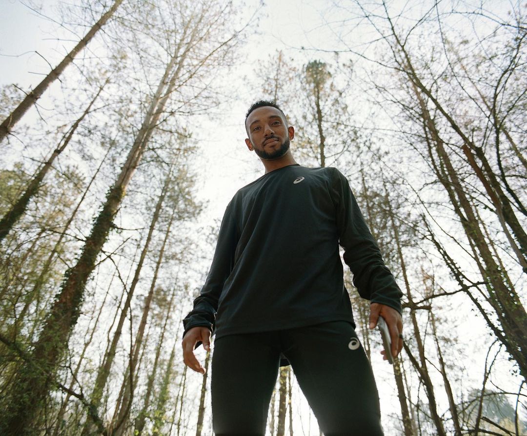 A man in sportswear gazes from a low angle, set against a forest backdrop, trees towering towards a bright sky, creating a dynamic view.