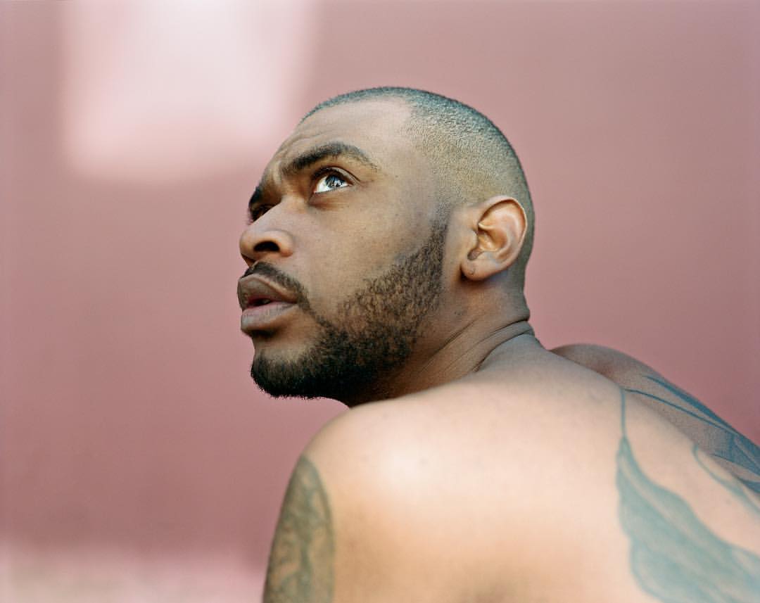 Low angle portrait of a Black adult male looking up and away from the camera, visible tattoos on the shoulder and arm.