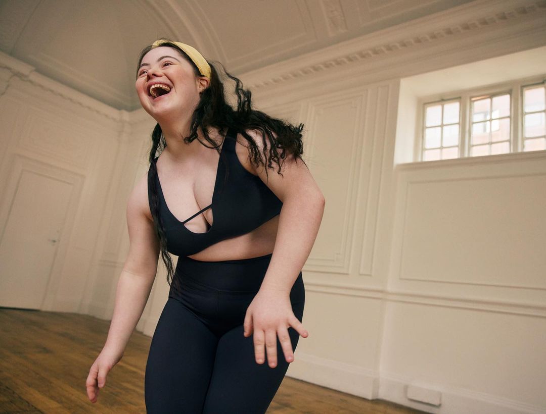 Laughing woman with Down syndrome wearing fitness attire stands in a bright room with architectural details. Joyful and authentic.