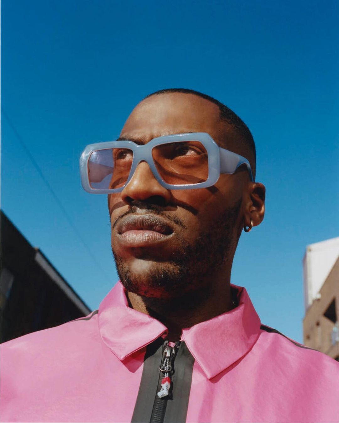 Close-up portrait of an African American man wearing light blue sunglasses and a pink jacket against a bright blue sky outdoors.