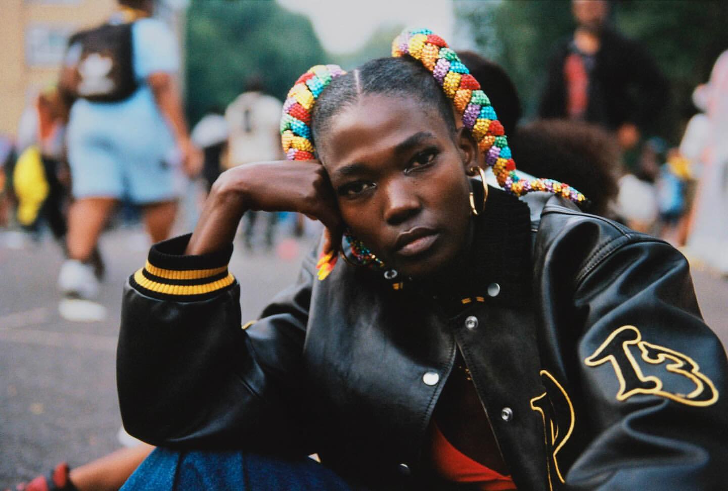 Stylish portrait of a Black woman with colorful braided pigtails, wearing a black jacket, sitting outdoors with blurred people in the background.