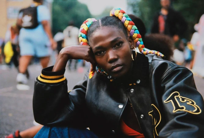 Stylish portrait of a Black woman with colorful braided pigtails, wearing a black jacket, sitting outdoors with blurred people in the background.