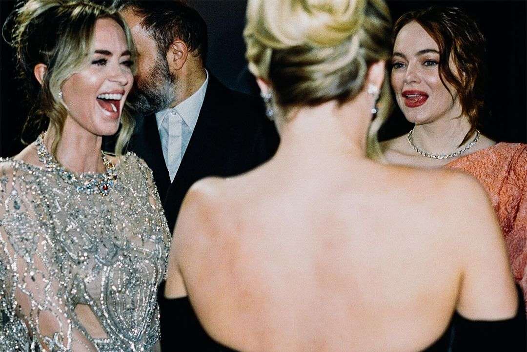 Candid shot of four people including actresses Emma Stone and Emily Blunt. Two women in evening gowns, one man in a suit in the background.