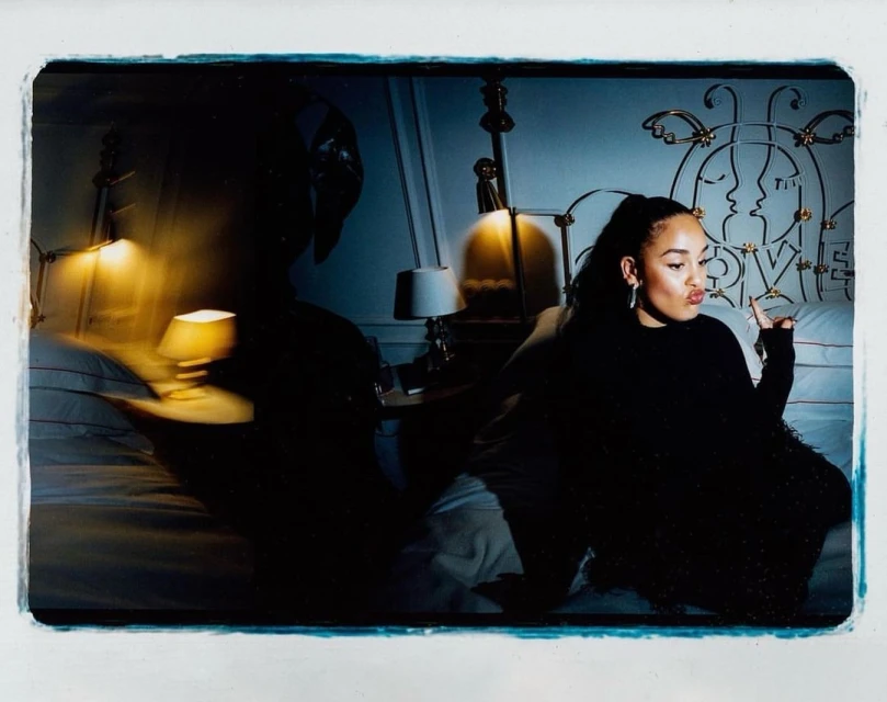 Indoor portrait of an adult female with a ponytail, wearing a dark top, seated on a bed in front of a decorative headboard and bedside lamps.