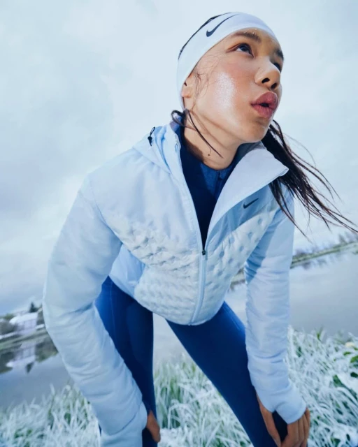An athletic woman wearing a Nike headband and jacket is outside in cool weather near a body of water, looking up and away.