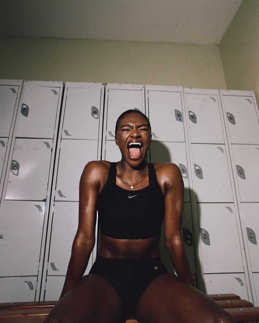 Sweaty female athlete with dark skin in a sports bra and shorts shouts while sitting in a locker room in front of metal lockers.
