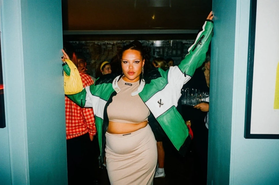 Woman in green and white jacket strikes a pose in doorway with partygoers in the background, holding a yellow purse.