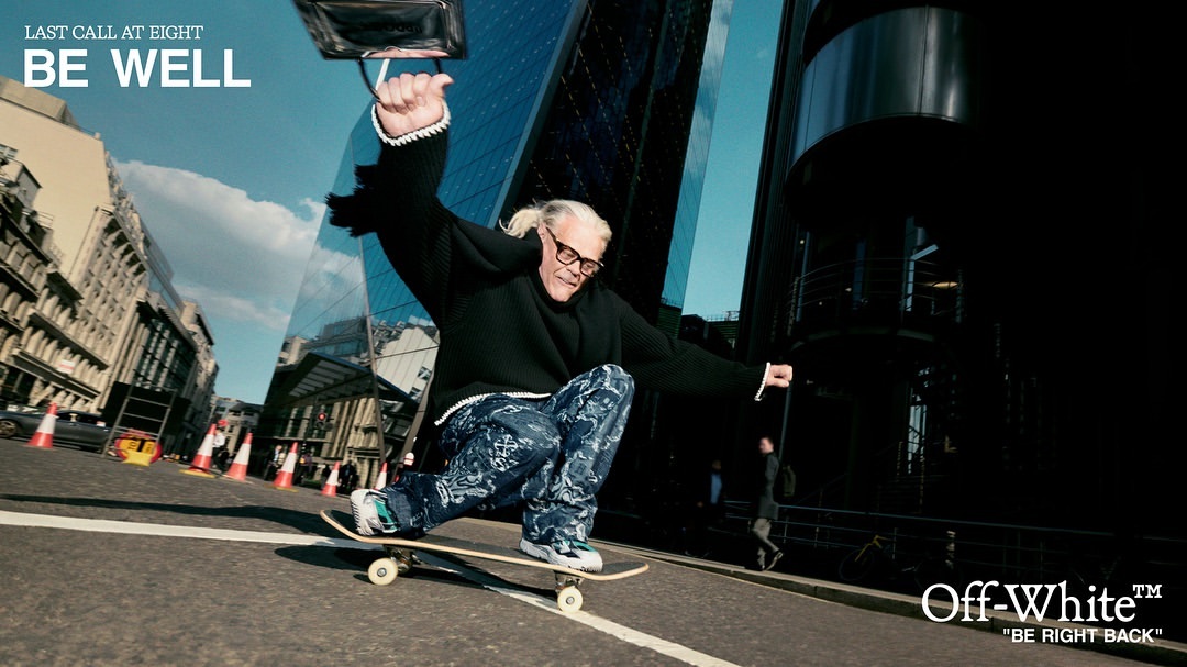 Elderly man in glasses and dark sweater rides skateboard down city street lined with buildings; text overlay reads 'Be Well' and 'Off-White'.