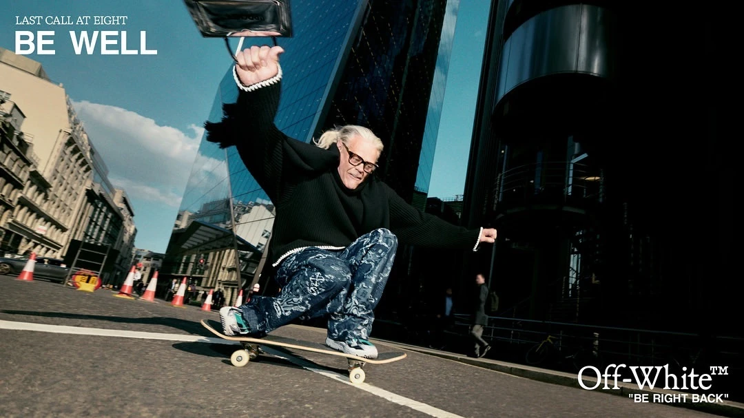 Elderly man in glasses and dark sweater rides skateboard down city street lined with buildings; text overlay reads 'Be Well' and 'Off-White'.