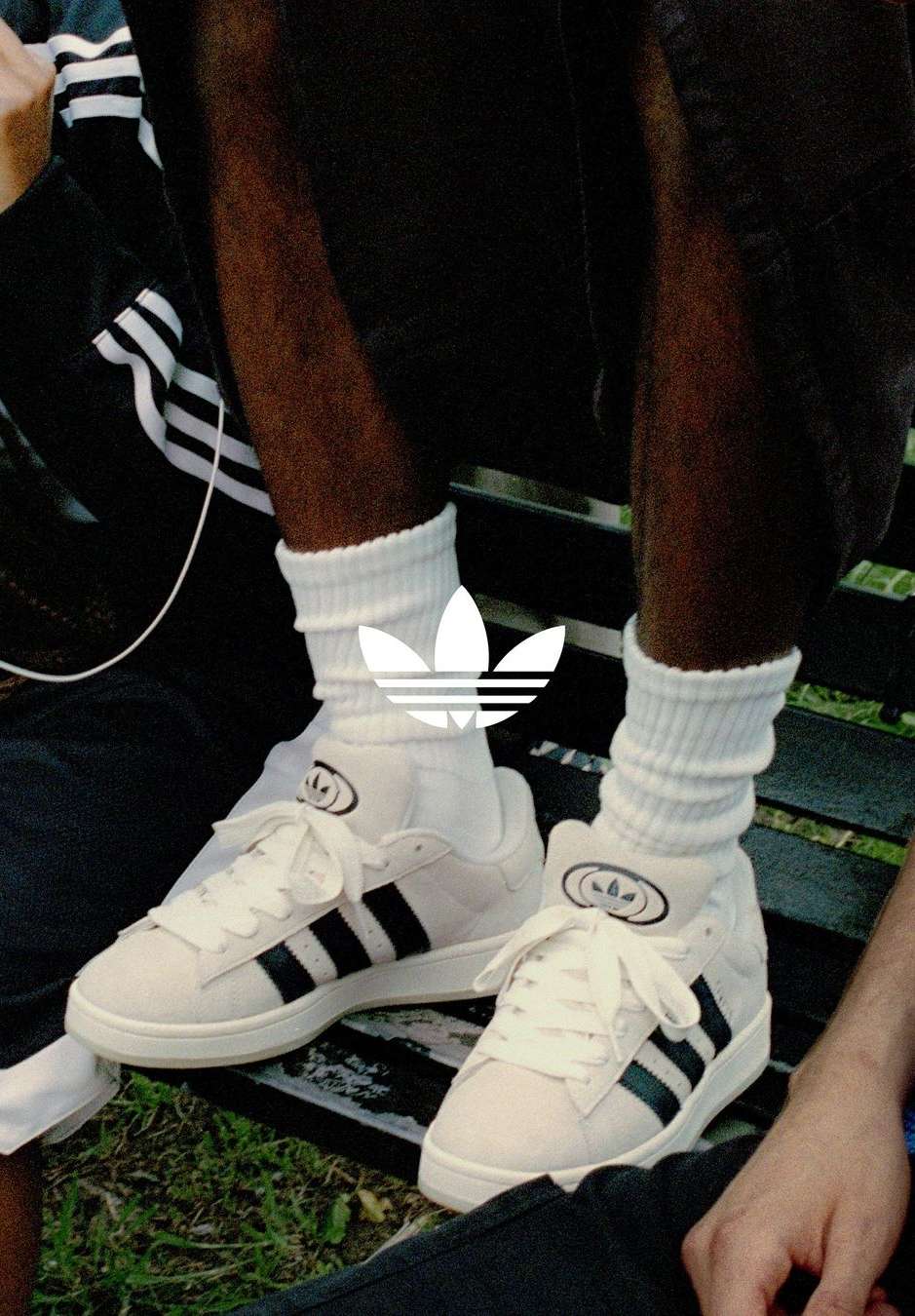Low angle of a person sitting on a bench in Adidas sneakers with black stripes and white socks, showcasing a mix of casual fashion and athletic style.