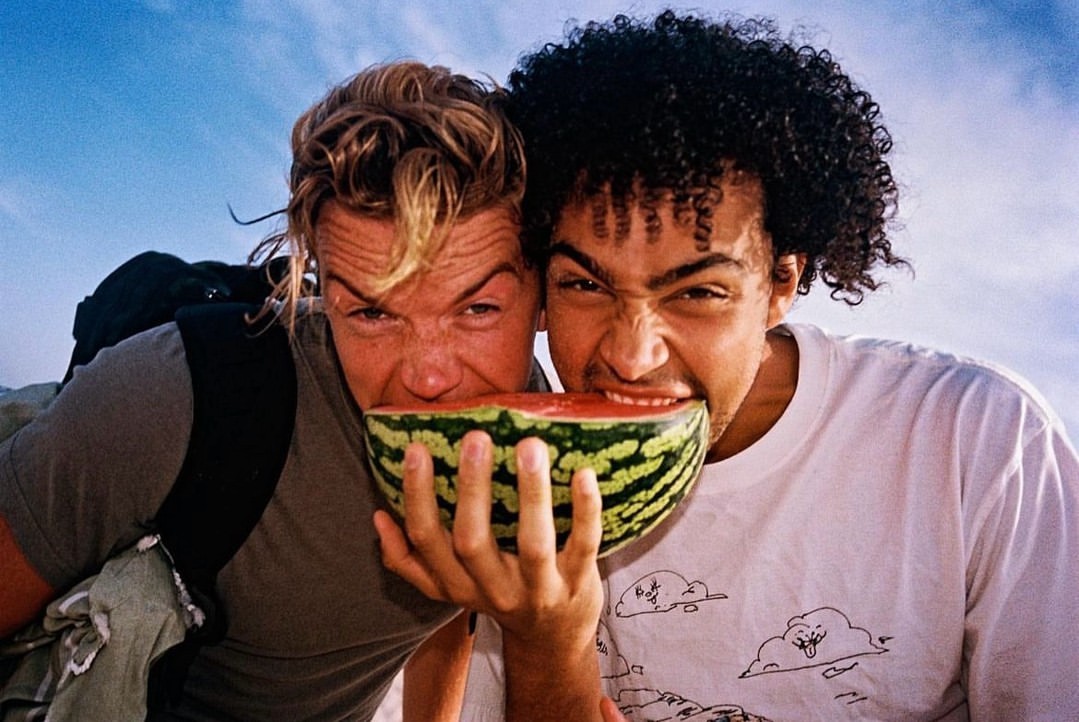 Two friends share a watermelon on a sunny day, smiling with the sky visible in the background, suggesting an outdoor, summery vibe.