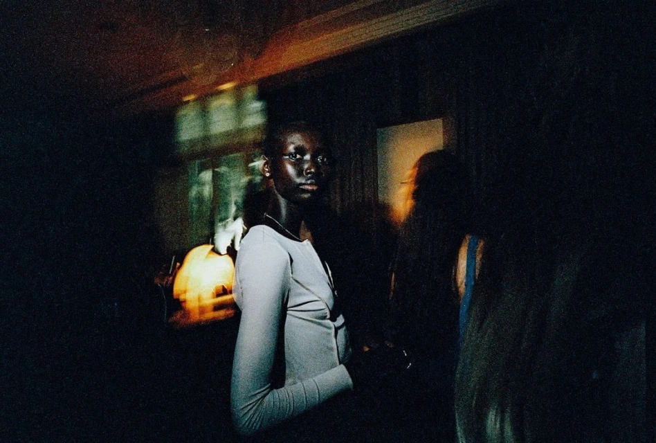 Portrait of an adult woman with dark skin wearing a light-gray long-sleeve top, standing indoors with soft lighting and a dark background.