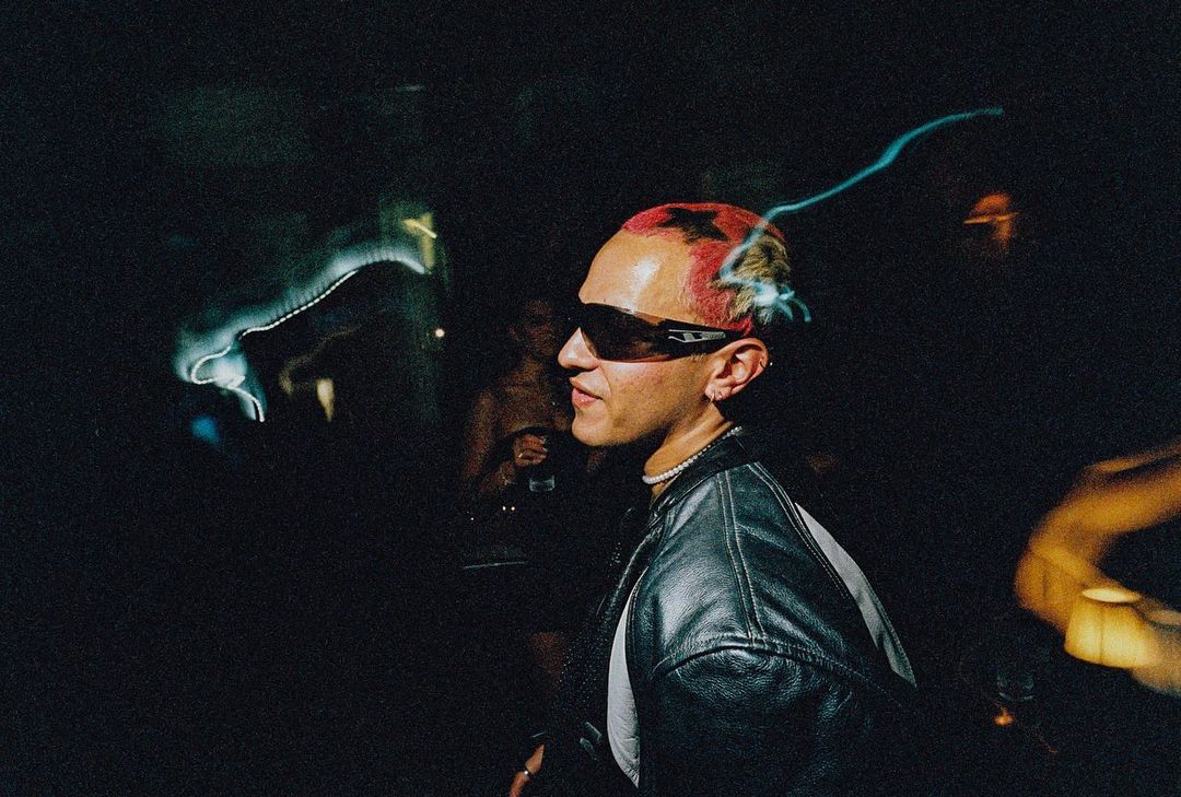 Person with dyed hair and sunglasses in dimly lit interior, wearing a black leather jacket, socializing at an event, close-up shot.