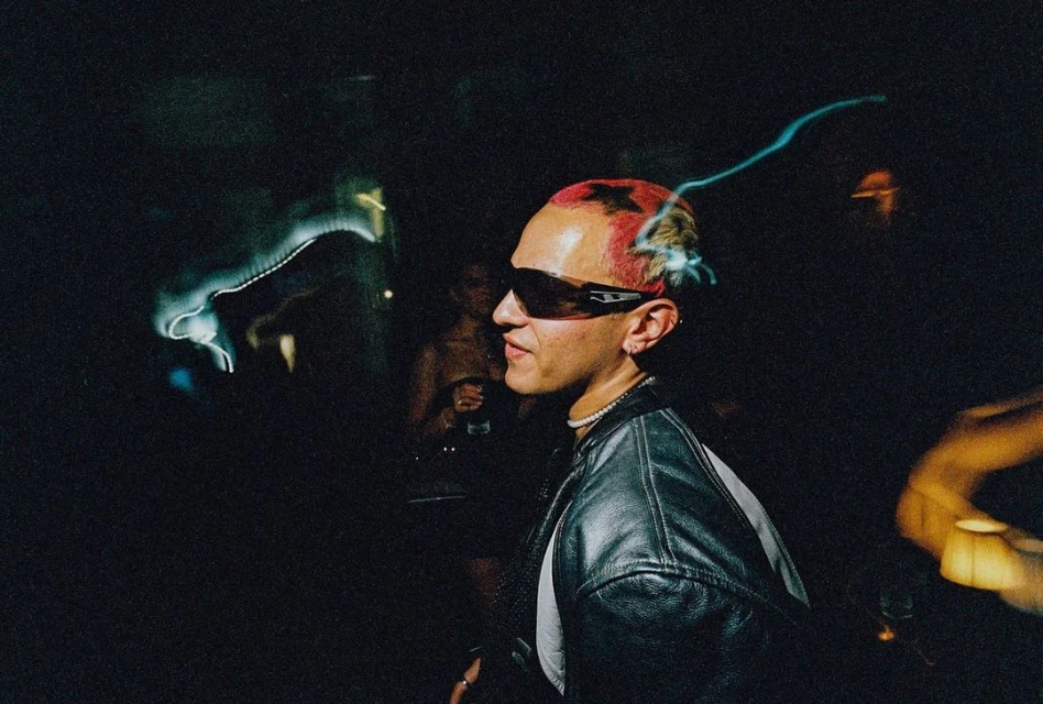 Person with dyed hair and sunglasses in dimly lit interior, wearing a black leather jacket, socializing at an event, close-up shot.