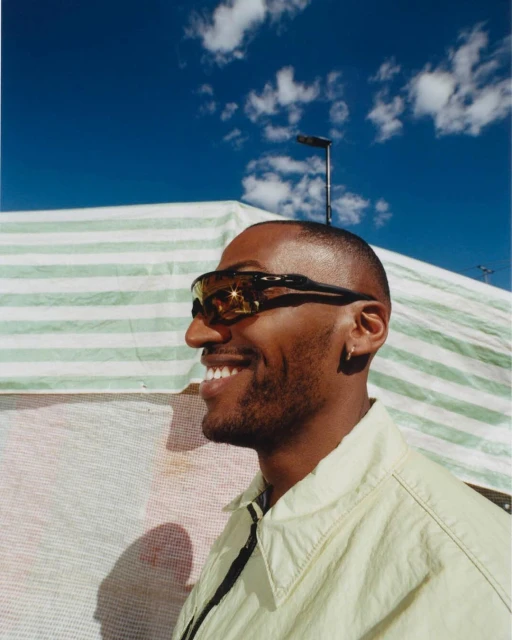 Close-up of a Black adult male in sunglasses, smiling with a light-yellow jacket against a striped awning and bright-blue sky.
