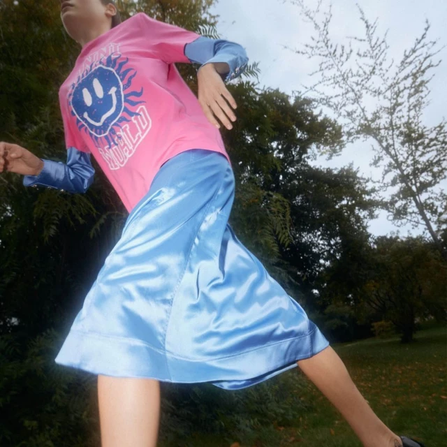 Adult in motion wearing a pink graphic tee and a blue skirt, posing outdoors against a backdrop of trees under an overcast sky.