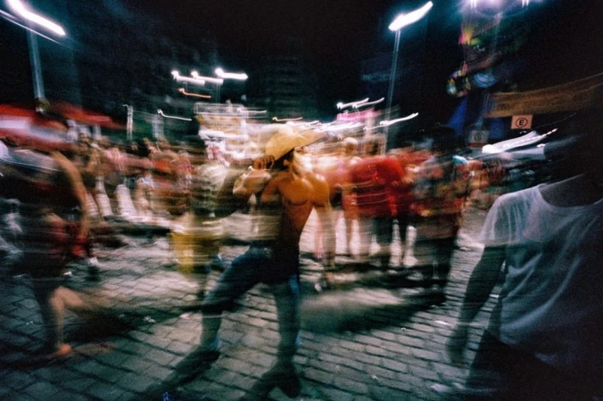 Nighttime street scene with a crowd of people, including a shirtless man in a hat, captured with motion blur and streetlights.