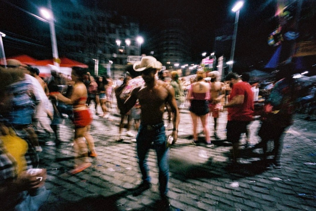 Nighttime urban crowd scene with shirtless man in jeans and a cowboy hat, street lights, and blurred motion suggesting lively activity.