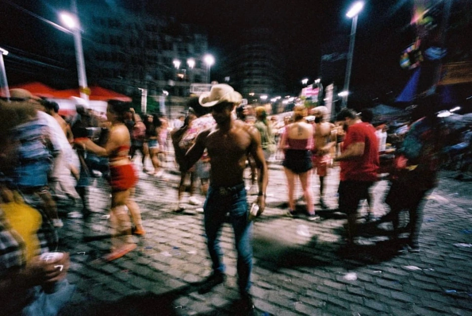 Nighttime urban crowd scene with shirtless man in jeans and a cowboy hat, street lights, and blurred motion suggesting lively activity.