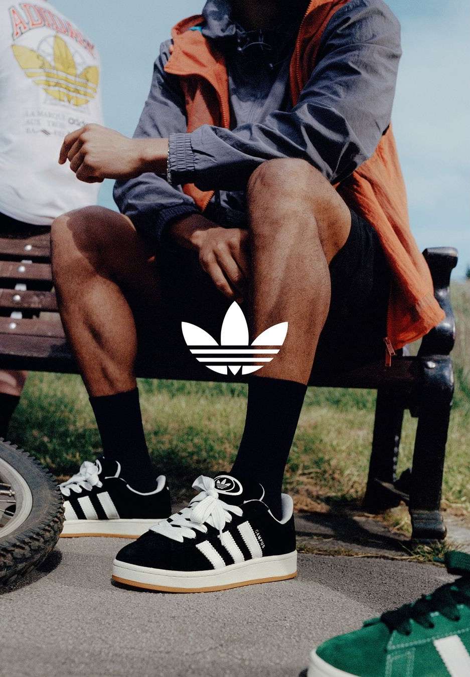 Three people sit on a bench in athletic wear, with focus on black and white Adidas sneakers, outside on a paved surface on a sunny day.