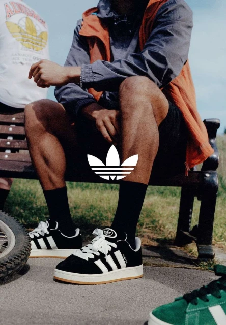Three people sit on a bench in athletic wear, with focus on black and white Adidas sneakers, outside on a paved surface on a sunny day.
