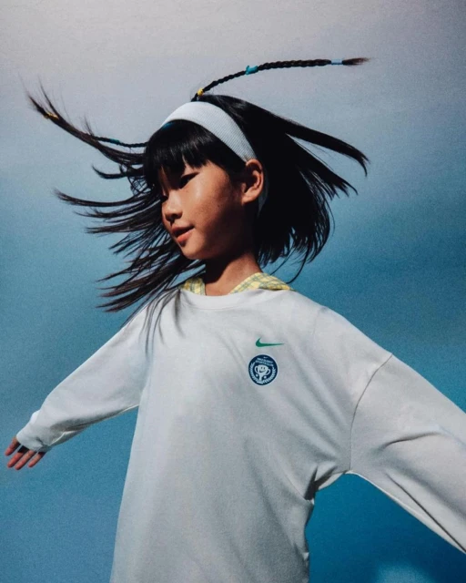 A young girl with dark hair and a white headband smiles against a light blue background. She wears a long-sleeved white shirt with a logo.