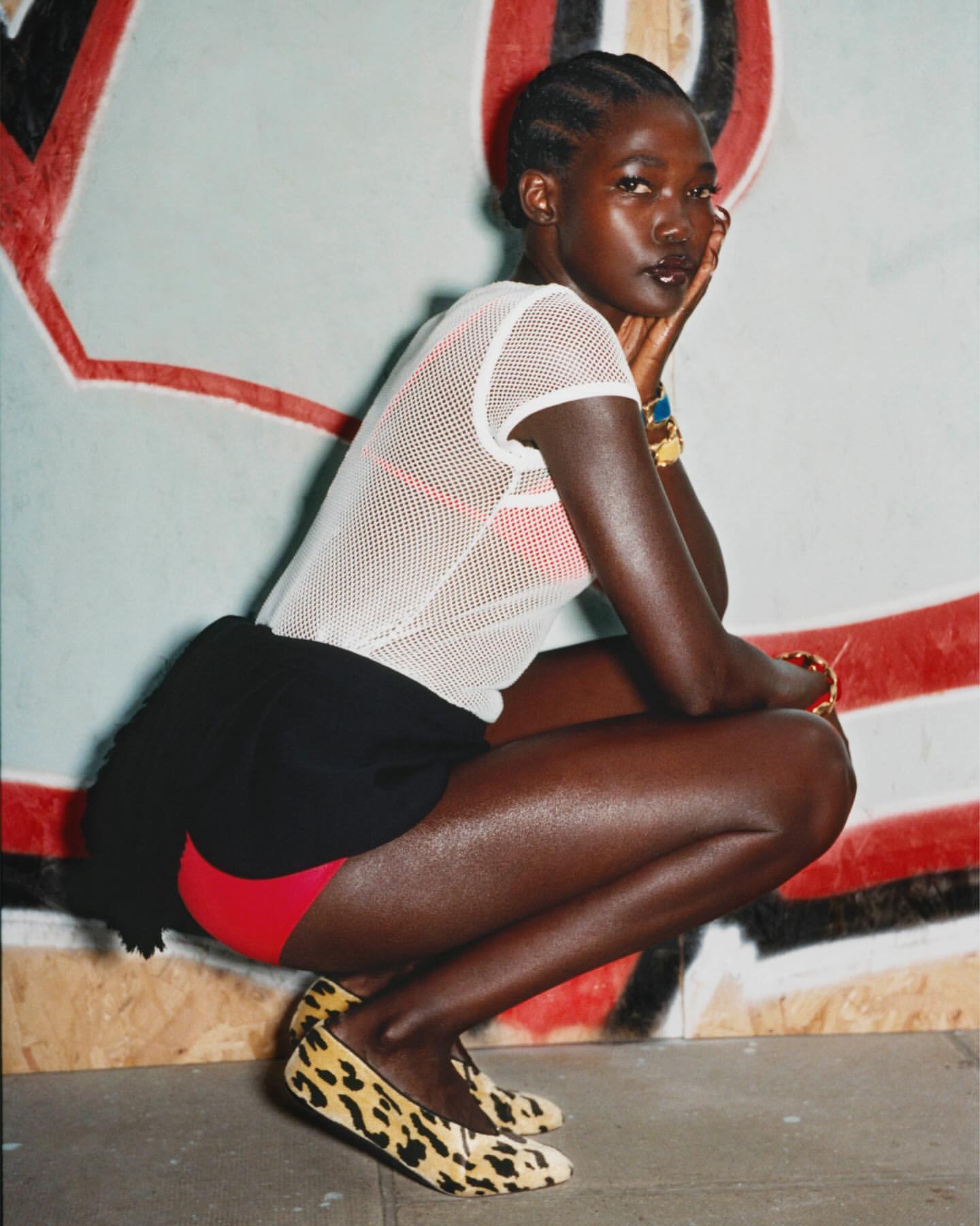 Adult female poses in front of a painted wall wearing a white mesh top, black shorts, red under shorts, and leopard print shoes.