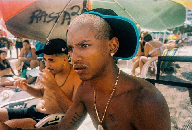 Portrait of shirtless adult male with buzzcut and face tattoo on crowded sandy beach, wearing a cap, gold chain, and earrings. 