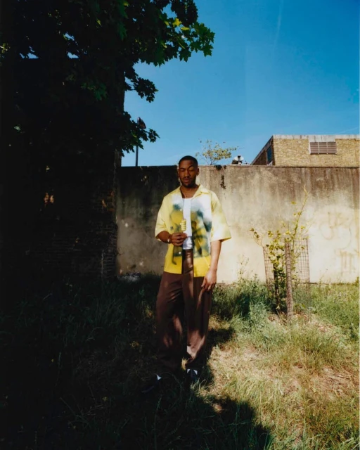 Full body shot of an African-American man standing on grass in a yellow shirt, brown pants, and white shoes under a blue sky.