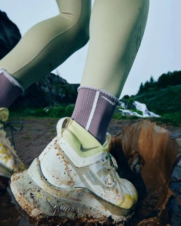 Close-up of a person's foot in a muddy white and yellow running shoe, stepping in a puddle on a trail with a waterfall in the background.