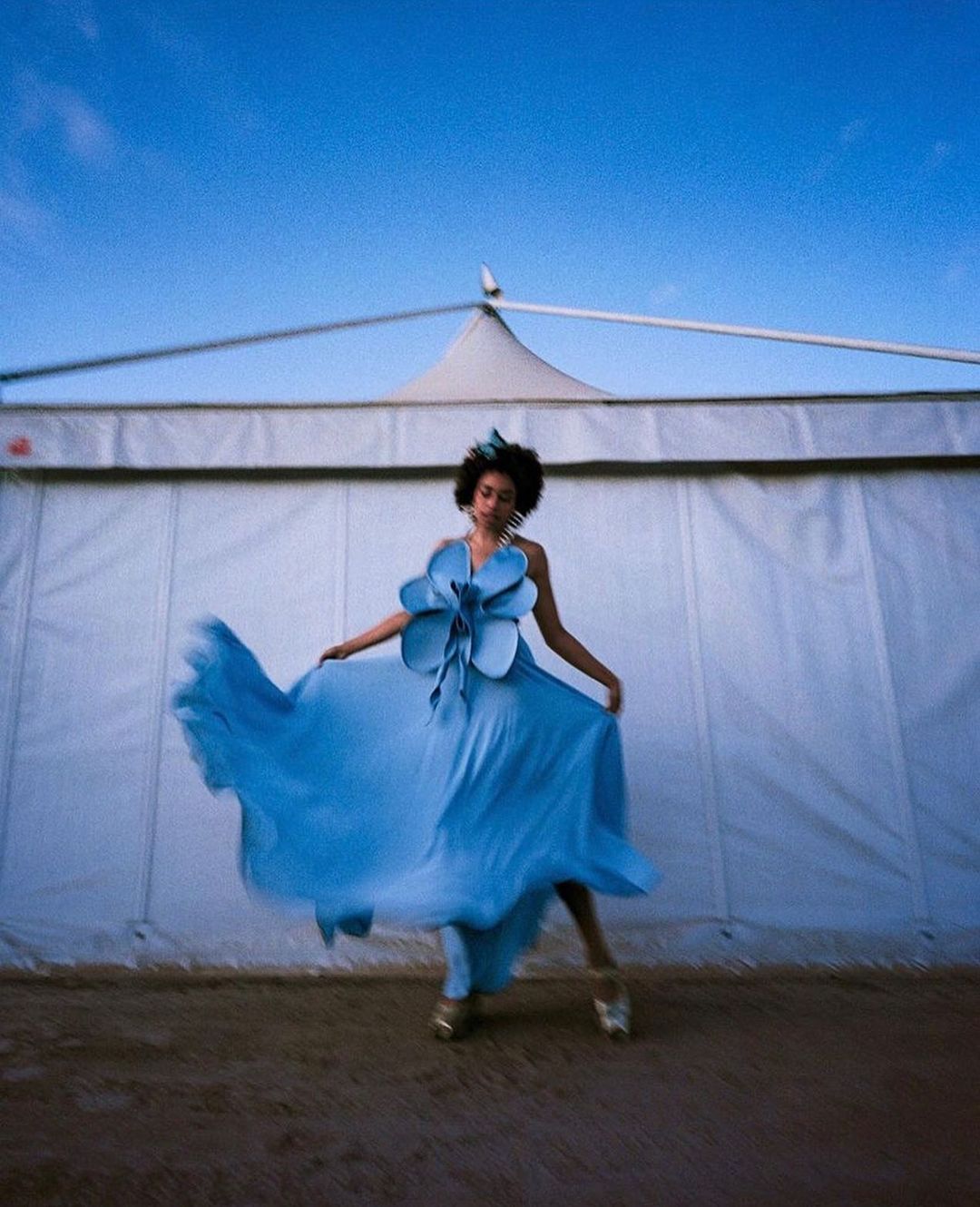 A woman in a blue couture dress poses outdoors in front of a white tent, set against a blue sky, showcasing fashion and artistic expression.