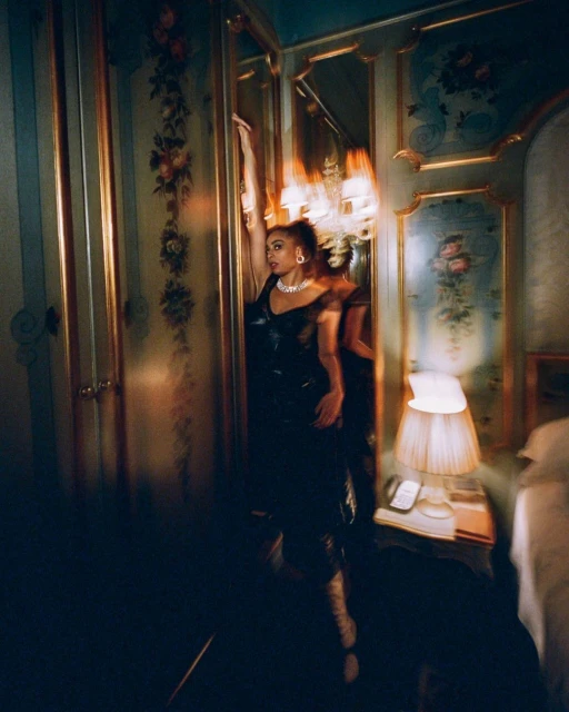 Adult female poses in black dress with raised arm inside an ornately decorated bedroom with floral wallpaper and mirrored closet.